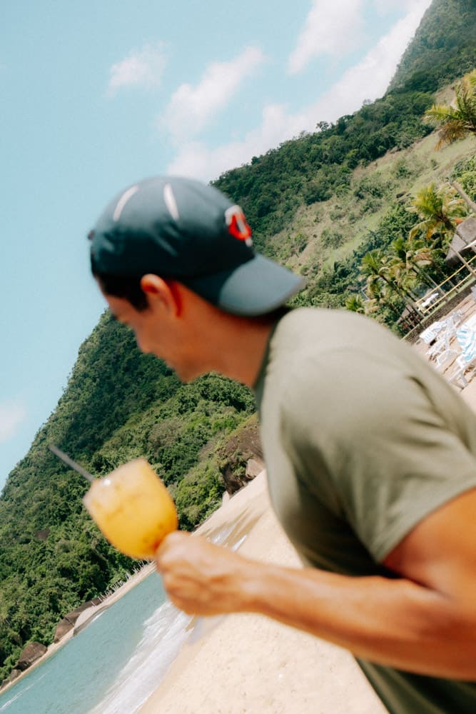 Homme en casquette tenant un spritz face aux montagnes, vacances au Club Med Rio das Pedras, Brésil