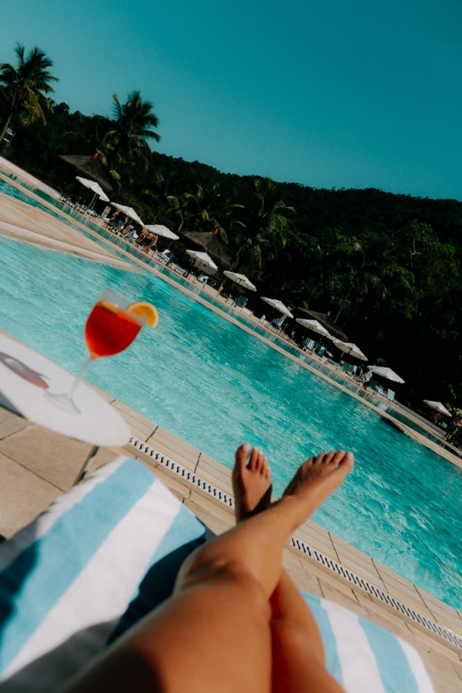 Jambes allongées sur un transat et spritz au bord de la piscine vus d'en haut, Club Med Rio das Pedras