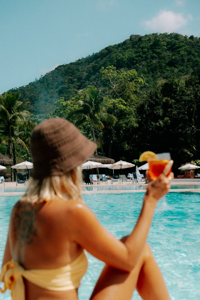 Femme en maillot jaune et bob assise au bord de la piscine, spritz à la main, Club Med Rio das Pedras
